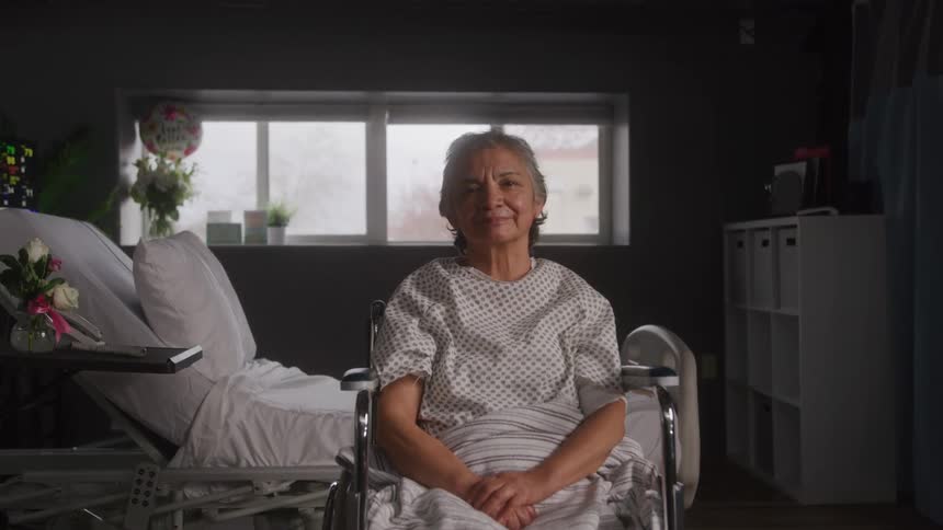 A woman with grey hair sits in a wheelchair in a hospital room looking directly at the camera with a small smile