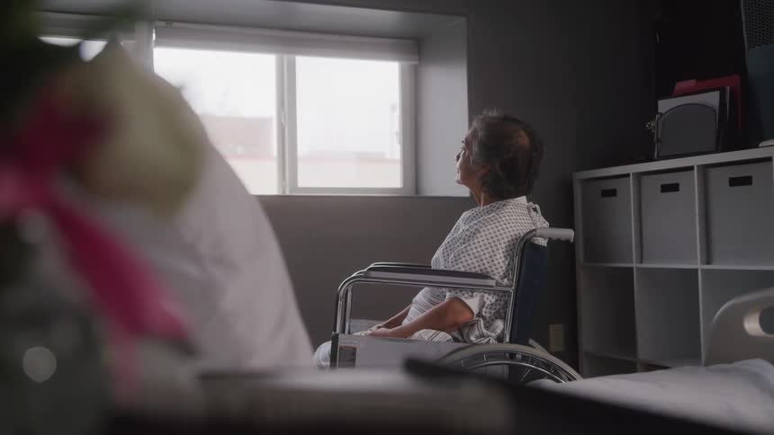 A woman with short grey hair sits in a wheelchair while looking out a large window in a hospital room