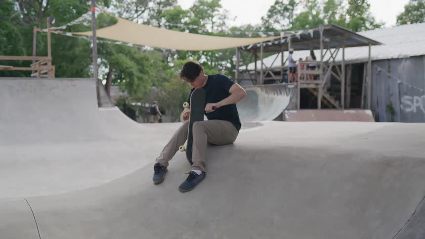 Young man sitting on the skate park holds his skateboard while looking down