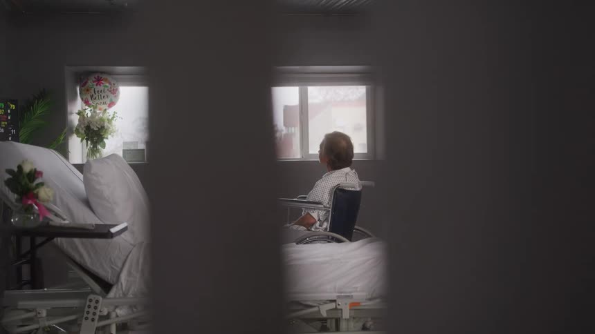 An elderly female patient sits on a wheelchair beside a bed in a hospital room during daytime