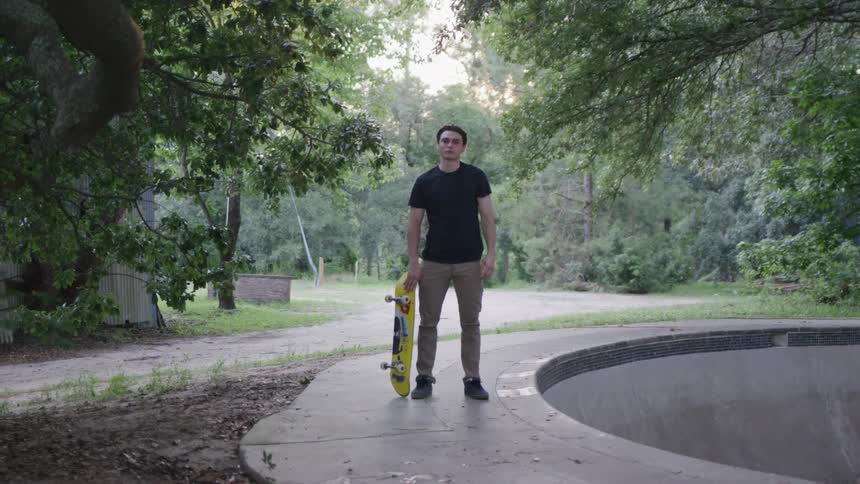 A man holding a skateboard stands in a skateboarding park surrounded by trees at midday