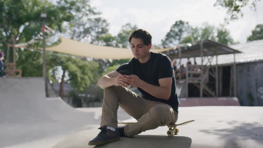 A man sits on a skateboard in a public park using his phone at midday