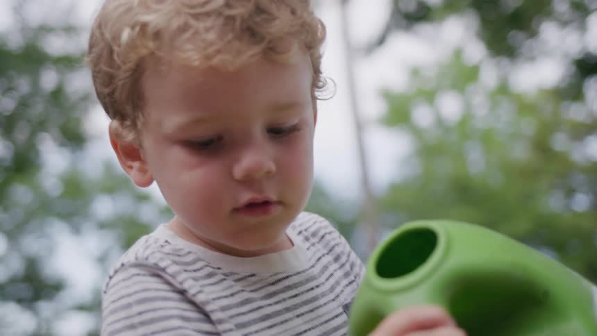 The video shows a closeup of a young child holding a green watering can the child with curly blonde hair is looking down at the watering can with a focused expression