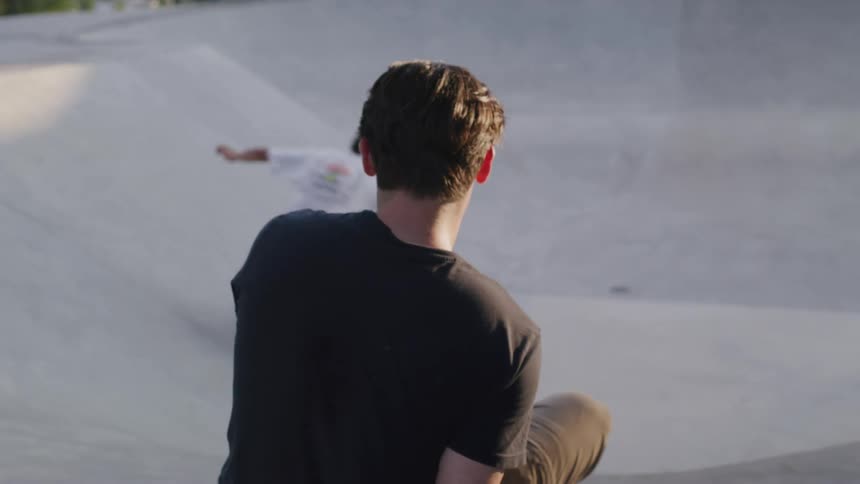 A man sitting at a skate park watching another man skateboarding and doing tricks