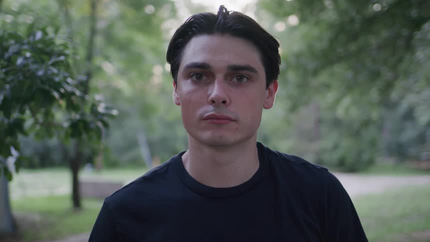 A young man with dark hair wearing a black tshirt stands in a slightly blurred outdoor setting and gazes thoughtfully into the camera
