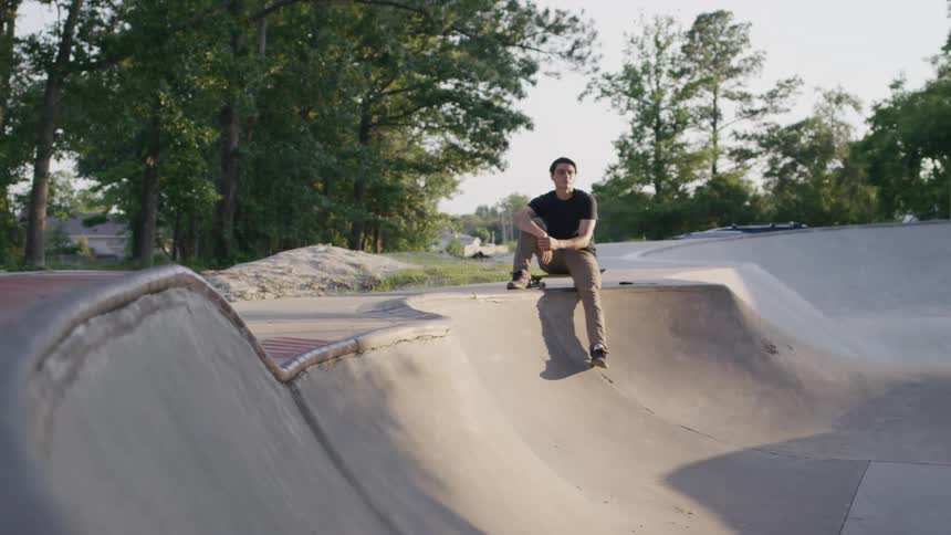 The video shows a young man sitting on the curved edge of a concrete skate bowl he is looking down and appears to be taking a break from skateboarding