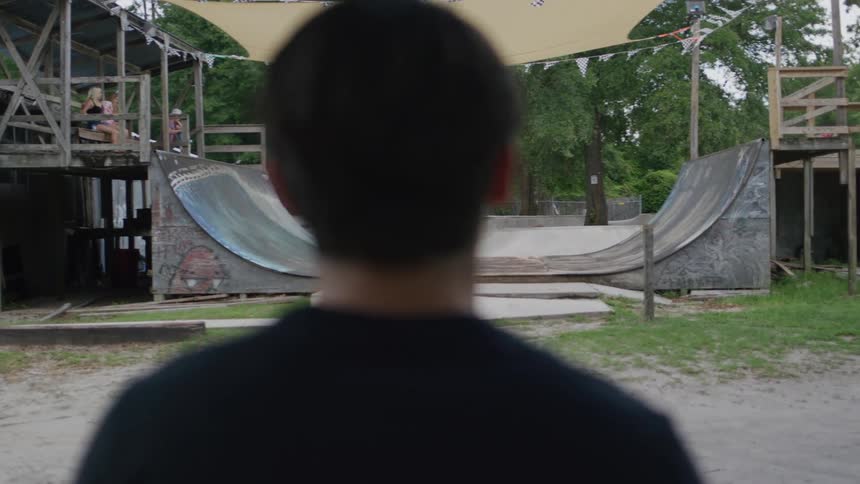 A male skateboarder wearing a beige shirt and a sun hat rides a skateboard up a large curved ramp at a skate park while a man watches in the foreground and two women and another man sit in the background