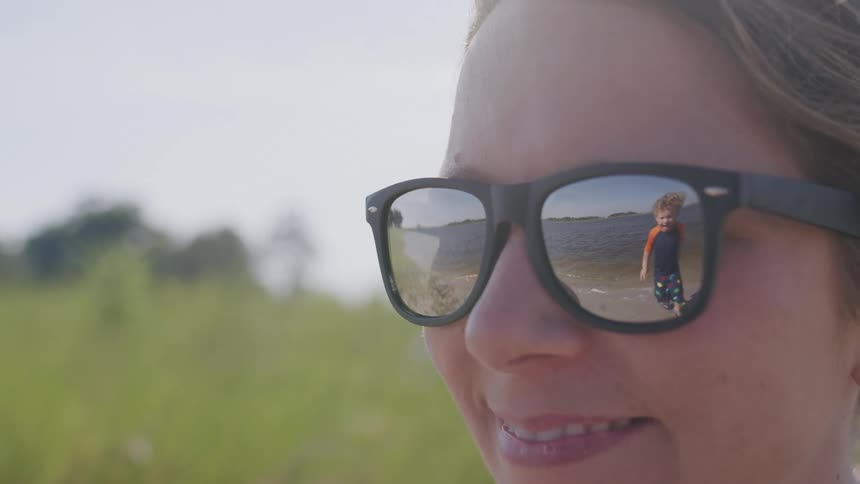 The video shows a closeup of a womans face with her sunglasses reflecting a small child running on the beach the woman is smiling and appears to be enjoying her time at the beach