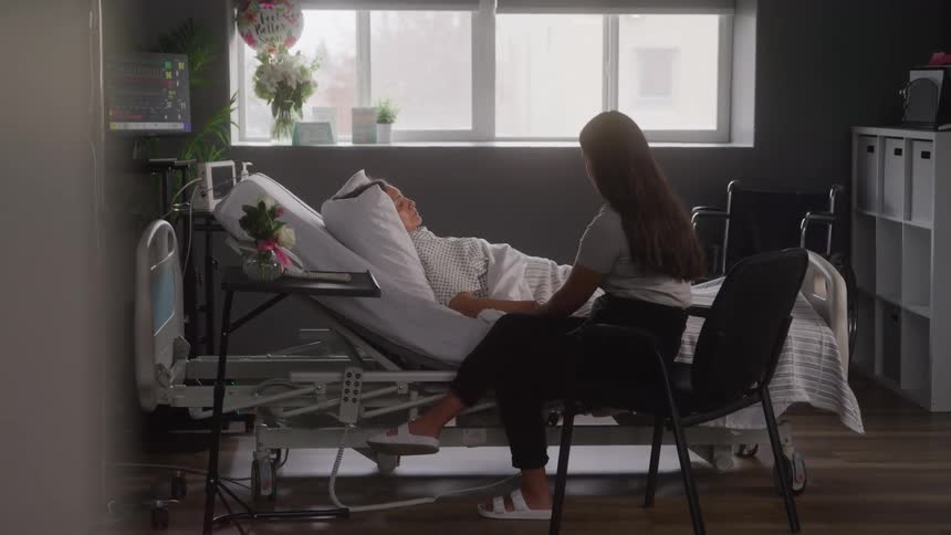 The video shows a hospital room with a patient lying in a bed while a young woman sits beside them in a chair the camera pans from left to right revealing the rooms details including a large window a bouquet of flowers and a heart monitor