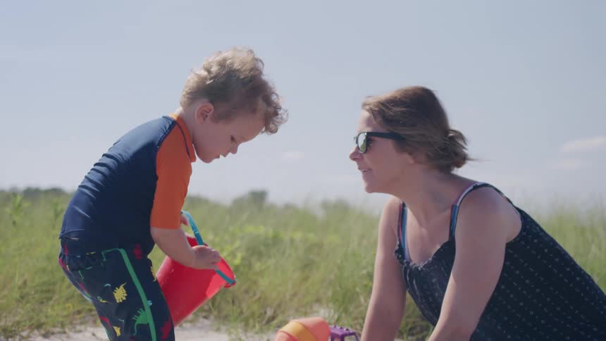 The video shows a mother and her young child playing with sand toys on a beach the child is wearing a blue and orange swim shirt and a colorful striped towel is laid out next to them