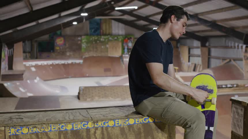 A focused male skateboarder sits in an indoor skate park intently rubbing the trucks of his yellow and purple skateboard to prepare it for use