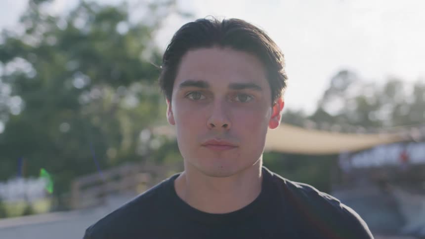 The video shows a closeup of a young man with dark hair looking directly at the camera he has a serious expression on his face and trees are blurred in the background
