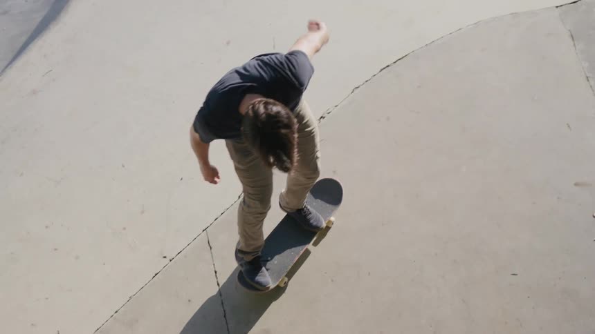 A man wearing a dark t shirt and light pants rides a skateboard on a curved concrete surface on a sunny day flipping the skateboard as he skates
