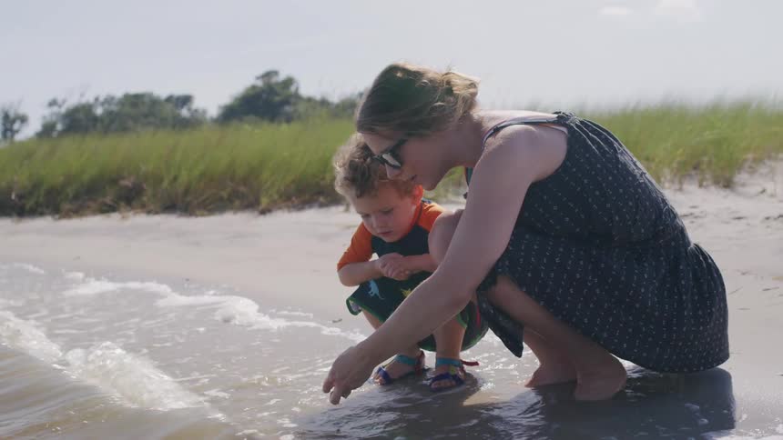 A woman in a dark sundress and a small boy in a patterned swim shirt crouch at the waters edge on a sandy beach playing with the water