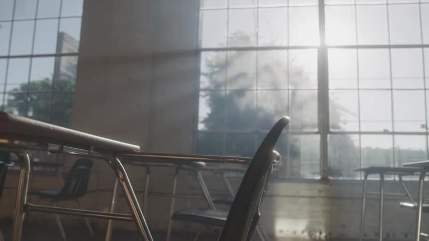 A beam of morning sunlight streams through tall classroom windows casting soft light across empty desks