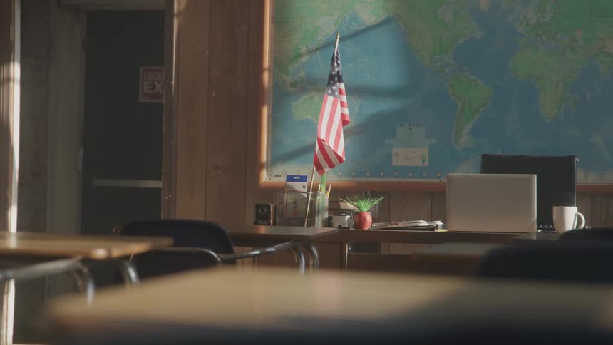 An american flag stands on a teachers desk with a laptop and a mug in front of a world map on a wood paneled wall while empty student desks are in the foreground