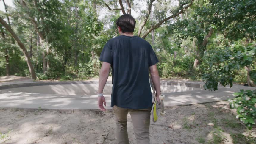 A young man wearing a dark tshirt and lightcolored pants walks away from the camera he is carrying a skateboard under his arm and walking toward a skatepark bowl