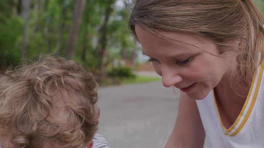 A woman with a ponytail and a small boy with curly hair crouch next to each other and look down at something outside of the frame