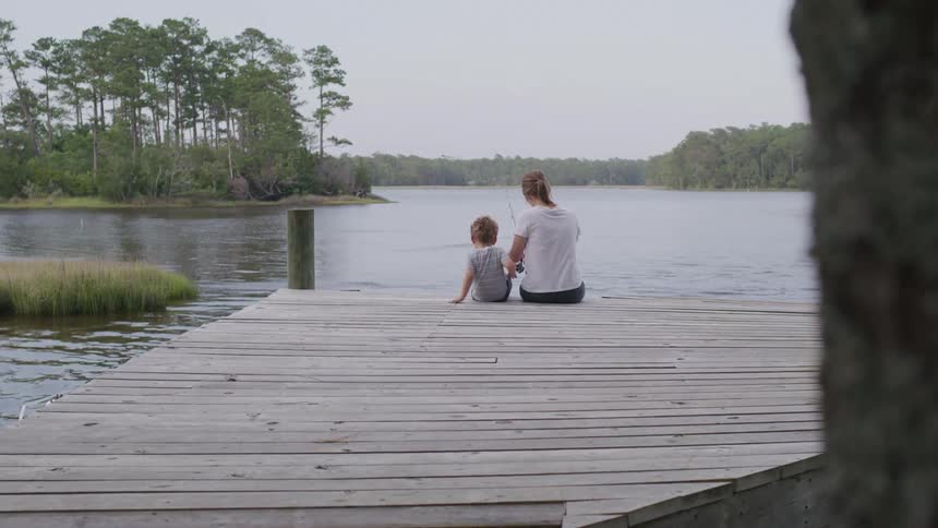 A woman sits at a dock with her son beside her fishing at midday