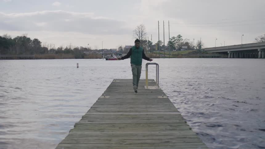 A man in a puffer vest jumps rope energetically along a dock over a calm river under a cloudy sky