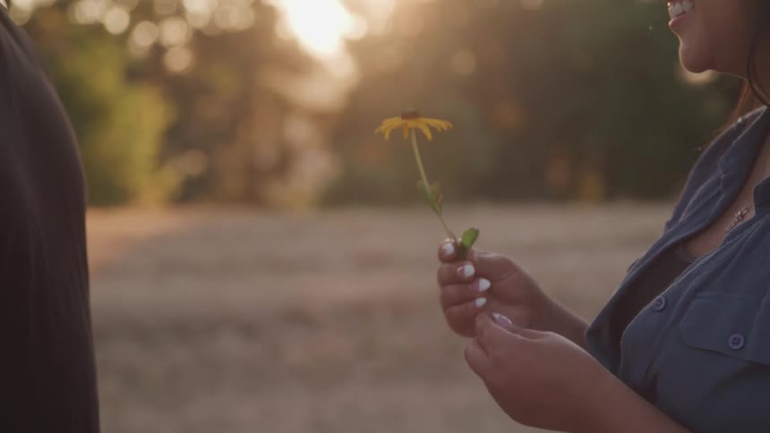 A man offers a single yellow flower to a woman outdoors as she leans in to take it and smell it