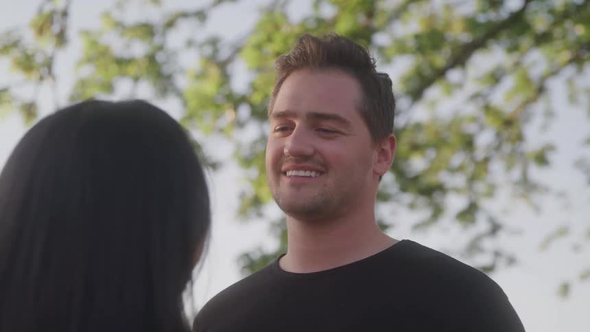 A man in a black shirt smiles warmly while standing near a woman outdoors beneath leafy trees