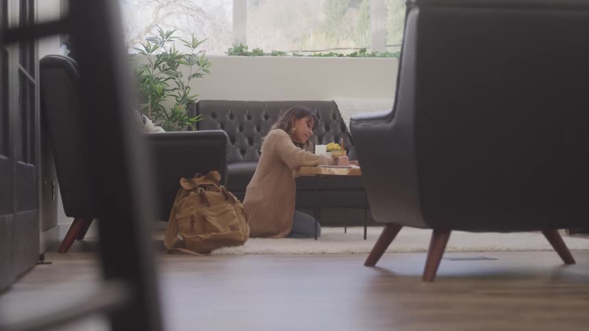 A young woman sits on the living room floor using the coffee table to do her homework wearing wired earbuds with a backpack resting beside an armchair at midday