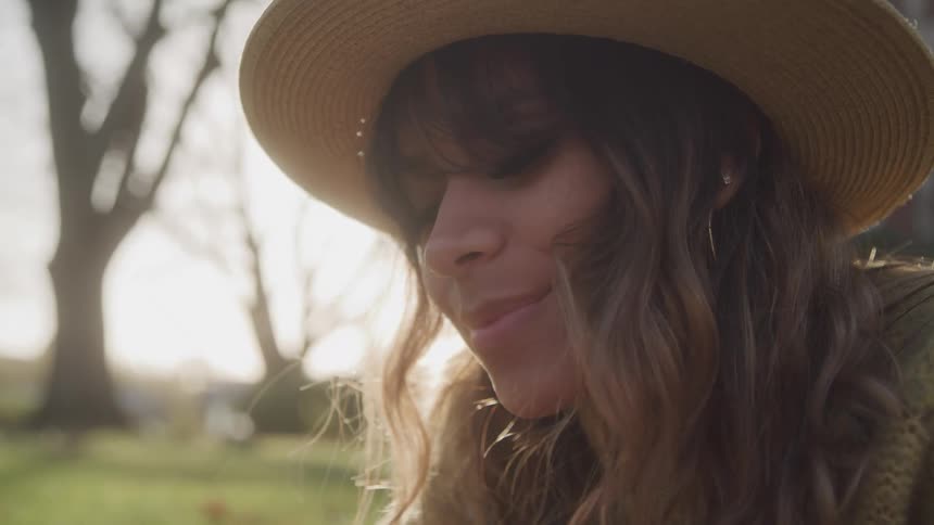 A woman wearing a tan straw hat smiles and looks down at an object outside of the frame with a sunlit background