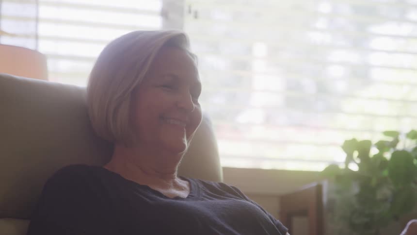 A woman waves and talks to another person during a video call on her phone while sitting on a chair in her living room at midday