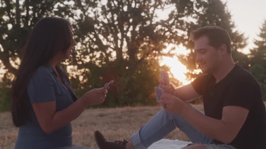 A couple plays cards while sitting on the grass during a picnic in a public park at sunset
