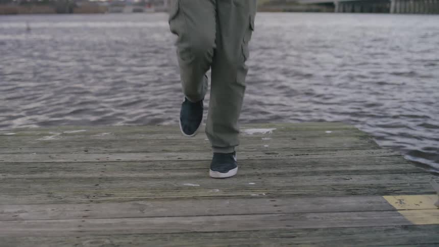 A male wearing sneakers jogs on a wooden dock beside a body of water is jumping rope