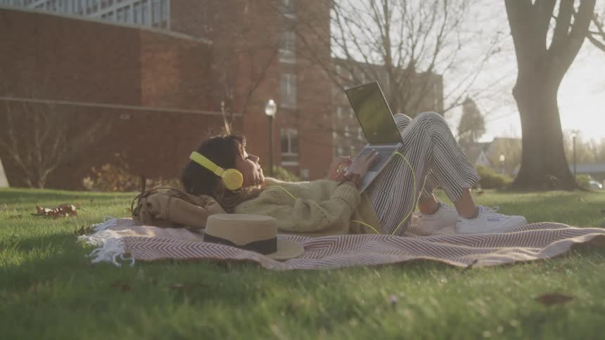 Woman lying on a blanket on the grass watches a laptop while wearing yellow headphones