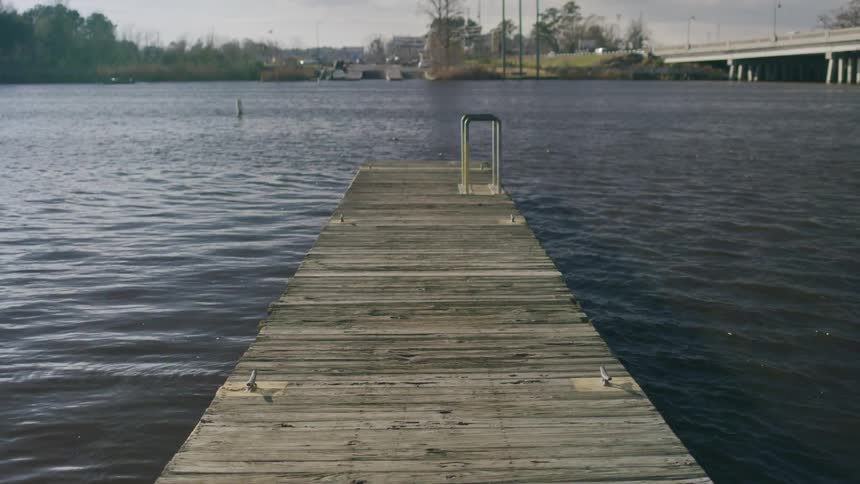 A wide shot shows a weathered wooden dock extending out into a wide body of water with a bridge and trees in the distance under a cloudy sky