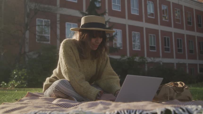A young woman sits on a lawn in front of a building wearing a hat and a green sweatshirt using her laptop under the sun