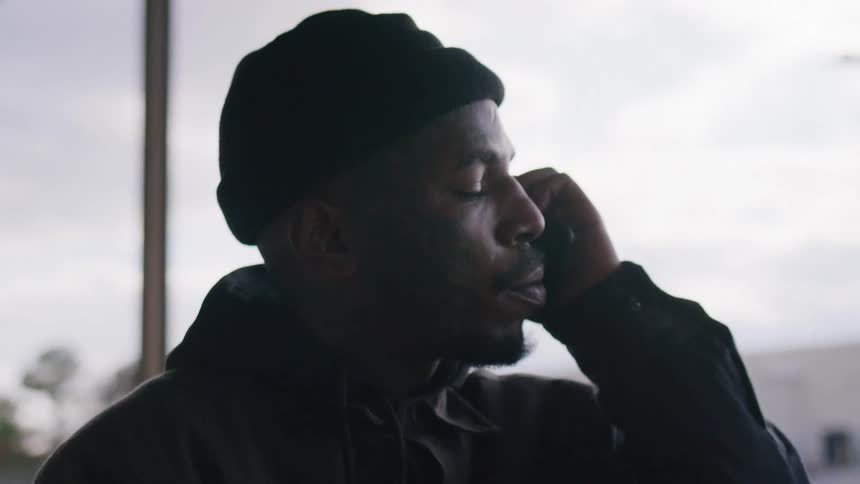 An angry black man dressed in dark clothing uses a smartphone outdoors under an overcast sky