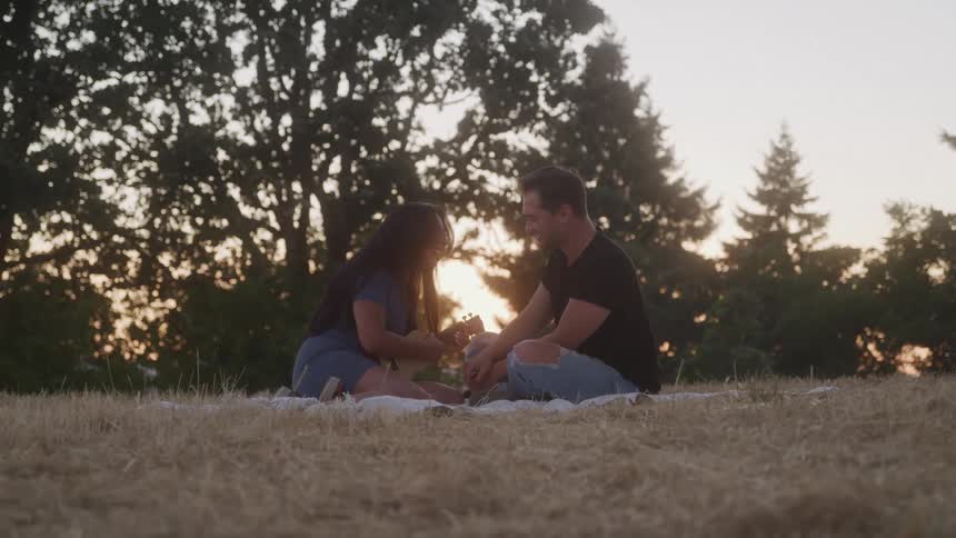 Couple sitting in a field at sunset while she plays ukulele