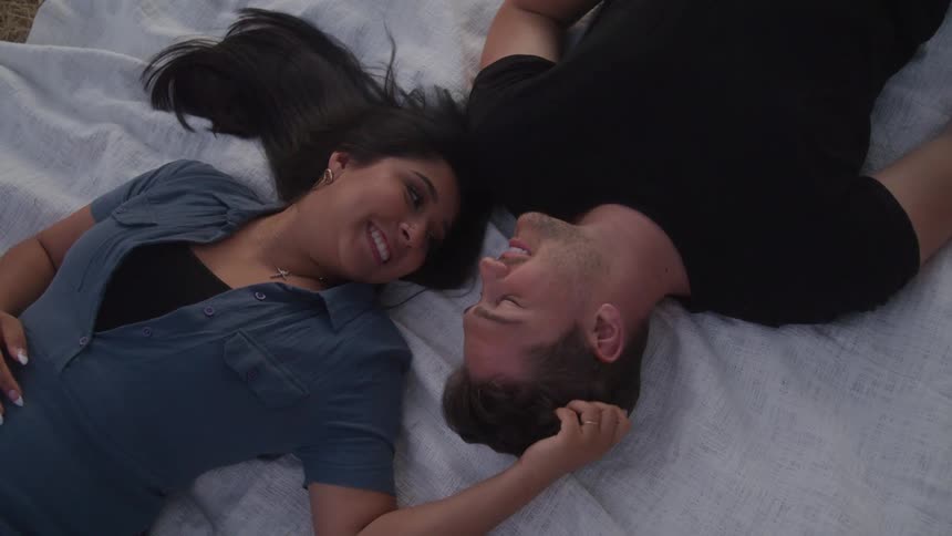 A man and a woman lie on a blanket on the ground looking and smiling at each other as the woman touches the mans hair