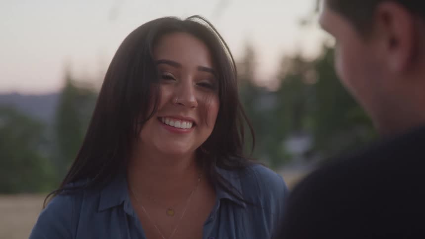 A woman with long dark hair and a nose ring smiles brightly while talking to a man outdoors at dusk