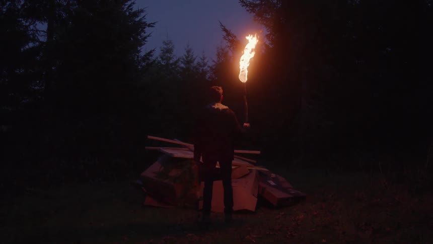 A man wearing a jacket and pants stands in a dark wooded area at night holding a flaming torch over a pile of cardboard and logs