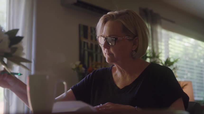 An older woman sitting at a table reading a book and holding a pen between her fingers