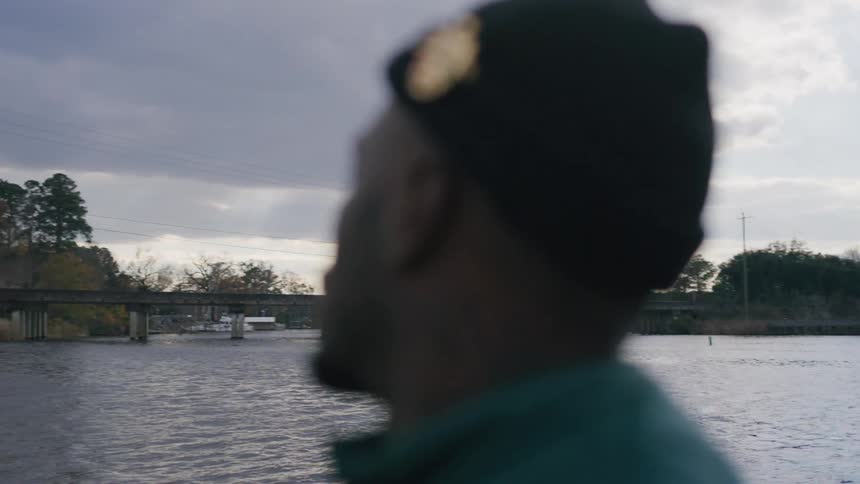 Man in a black beanie and green jacket looks thoughtful near a river