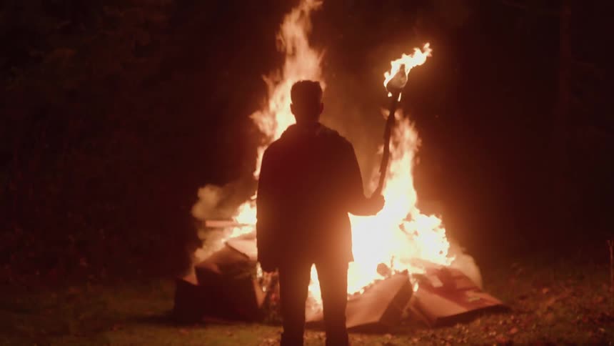 Silhouette of a man holding a torch in front of a campfire in a forest at night