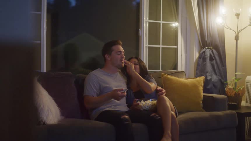 A man and a woman sit on a couch in a dark room with a window eating popcorn from a bowl while watching something together