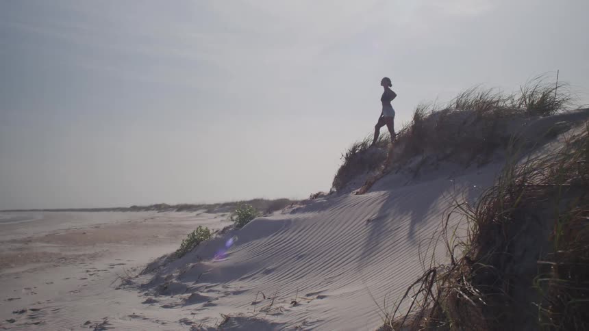 A person stands on top of a sandy dune covered in tall grass looking out at a wide empty beach on a bright day