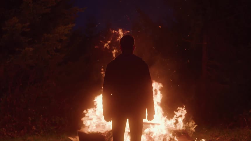 Man from behind watches a large bonfire in the darkness