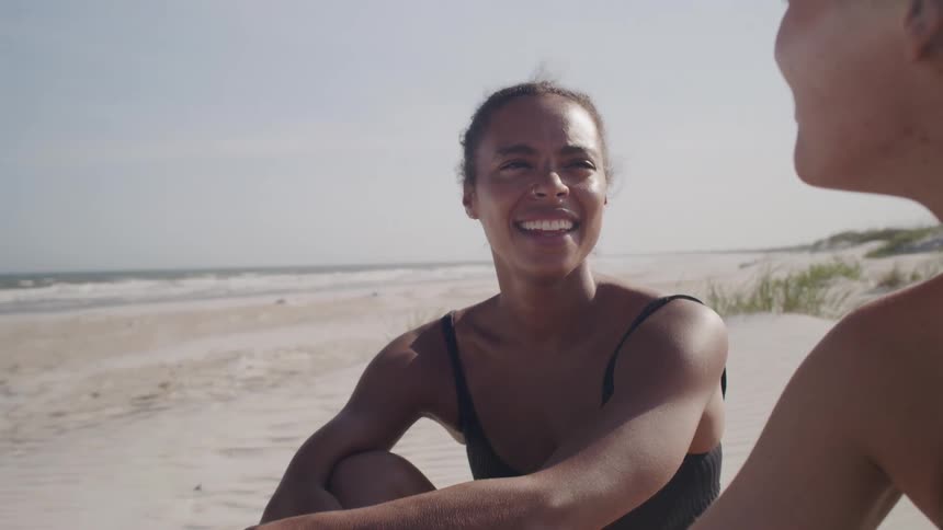 Two women in swimsuits talk and smile while sitting on the beach sand