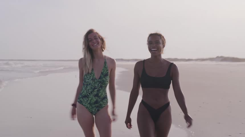 Two women in swimsuits walk smiling along the shoreline