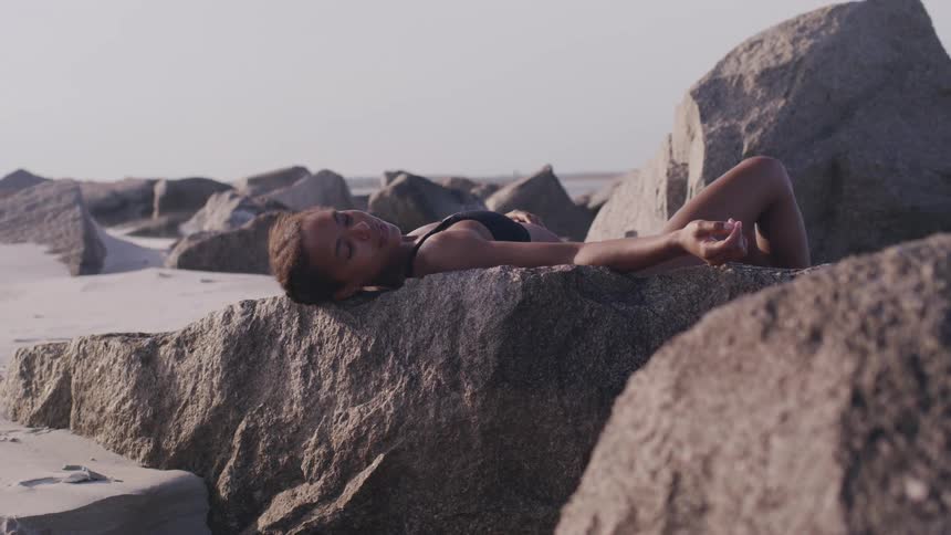 A woman in a black swimsuit reclines peacefully on large sunlit rocks at the beach