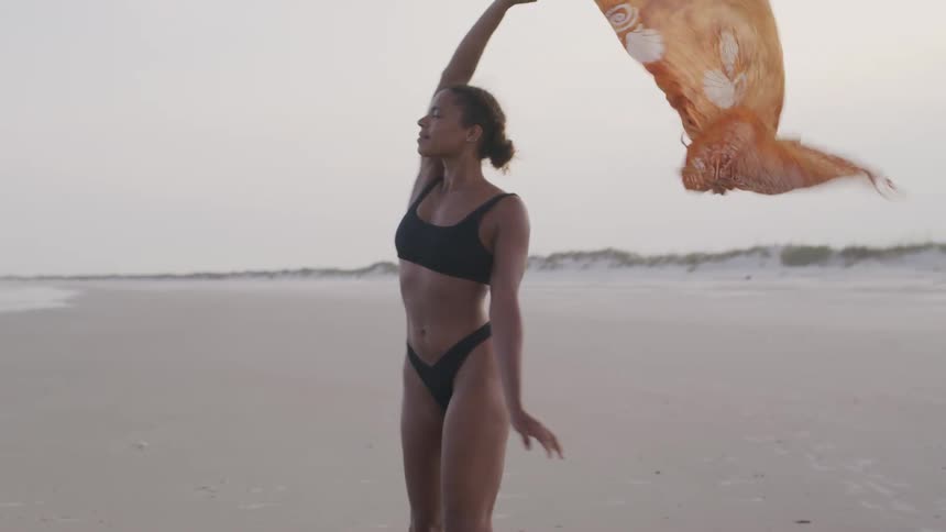 A woman in a black bikini is walking on a sandy beach she is holding a yellow scarf which is fluttering in the wind behind her