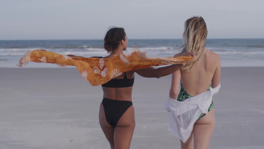 Two young women are shown from behind walking on the beach